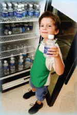 Boy holding a bottle of water standing in front of an open refrigerator filled with bottled water